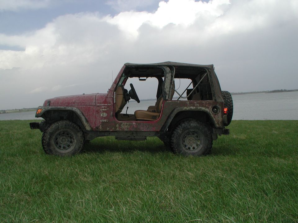 Red Jeep Wrangler with soft top removed, parked on a grassy field near a body of water