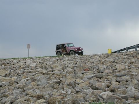 1998 Jeep TJ - A red Jeep Wrangler is parked on a rocky slope, with a yellow barrel and guardrail visible in the background.