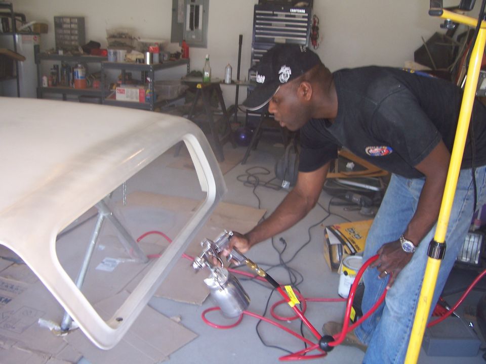 Man in garage sprays paint onto a detached vehicle hardtop. Tools and shelves are visible in the background