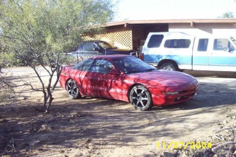 1995 Ford Probe - A red coupe with aftermarket wheels is parked on a dirt lot next to a blue and white pickup truck.