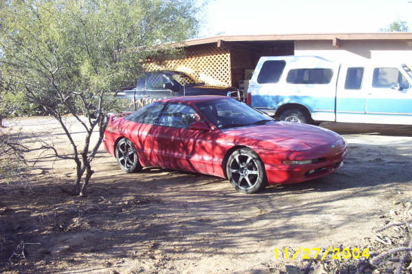 Red coupe with aftermarket wheels parked on dirt, a blue and white pickup truck behind it