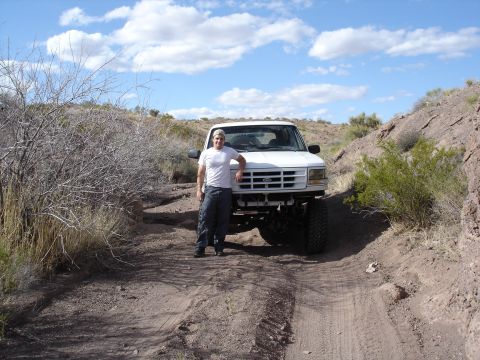 1996 Ford Bronco - A man stands next to a white Ford Bronco on a dusty, off-road trail under a cloudy sky.