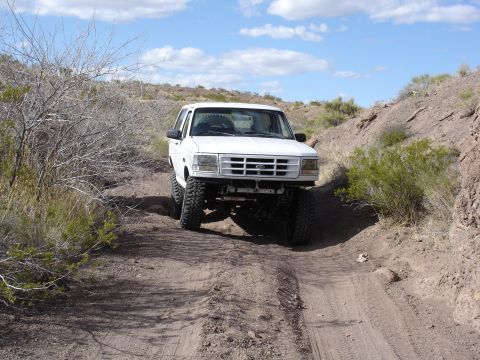1996 Ford Bronco - A white 1996 Ford Bronco navigates a dusty, off-road trail under a partly cloudy sky.