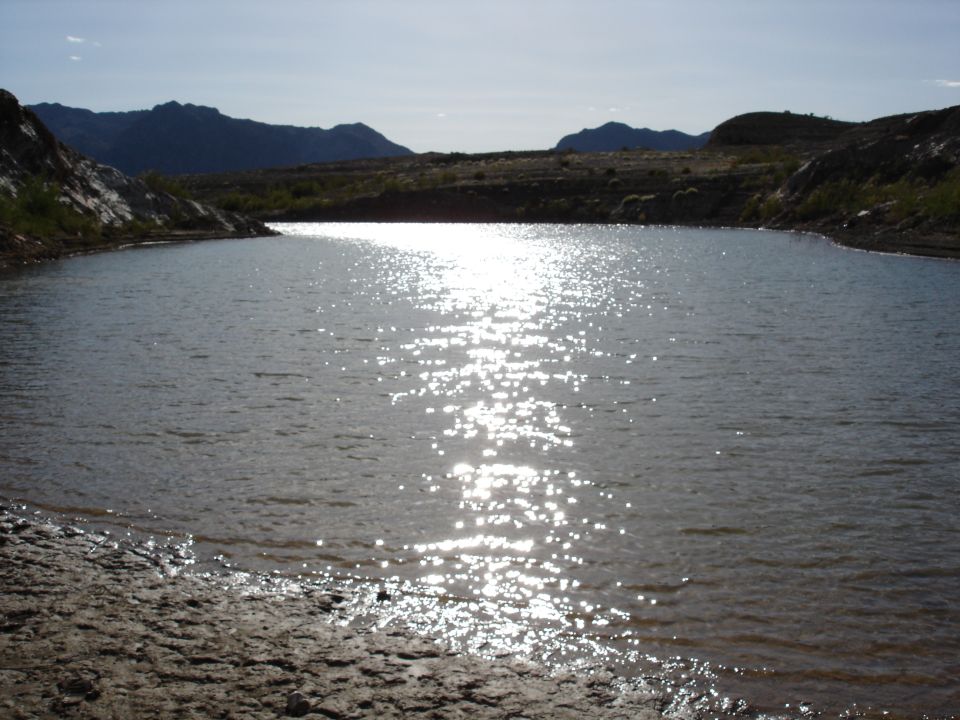 Water reflects sunlight under a bright sky with distant mountains and rocky shore