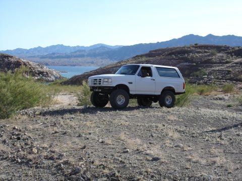 1996 Ford Bronco - A lifted 1996 Ford Bronco sits on a rocky incline with a lake and mountains in the background.