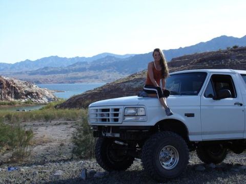 1996 Ford Bronco - Woman sits on the hood of a 1996 Ford Bronco parked in a desert landscape near a lake.