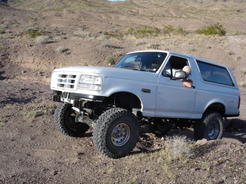 1996 Ford Bronco - A white 1996 Ford Bronco navigates a rocky desert terrain with a lifted suspension.