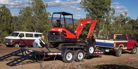 1990 Ford Bronco - A red pickup truck tows a trailer carrying a Kubota excavator, with a van visible in the background.