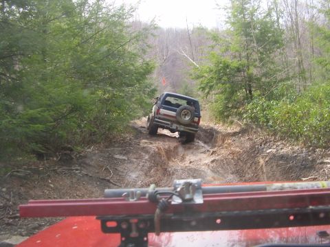 2003 Jeep Wrangler - A blue SUV navigates a challenging muddy trail, with the view from another vehicle in the foreground.