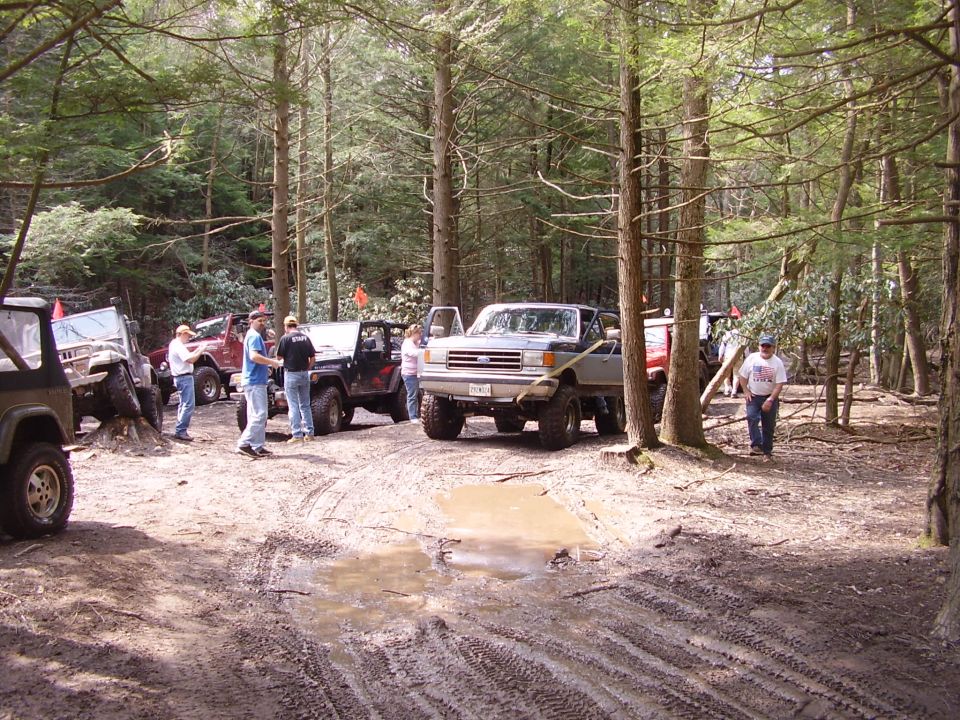 Several off-road vehicles, including Jeeps and a pickup truck, are parked on a muddy trail in a forest. People are standing around the vehicles