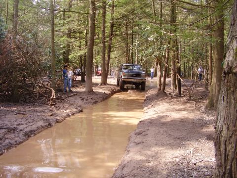 2003 Jeep Wrangler - A 1992 Ford F-150 navigates a muddy water obstacle in a wooded area, with spectators watching.
