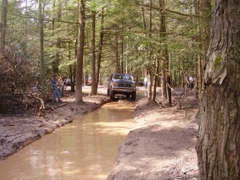 2003 Jeep Wrangler - Ford F-Series truck navigates a muddy trail in a forest with onlookers present.