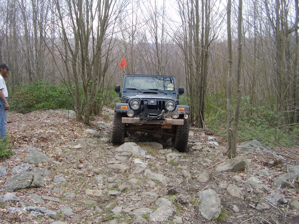 Jeep Wrangler driving on a rocky path in the woods. A person stands to the left