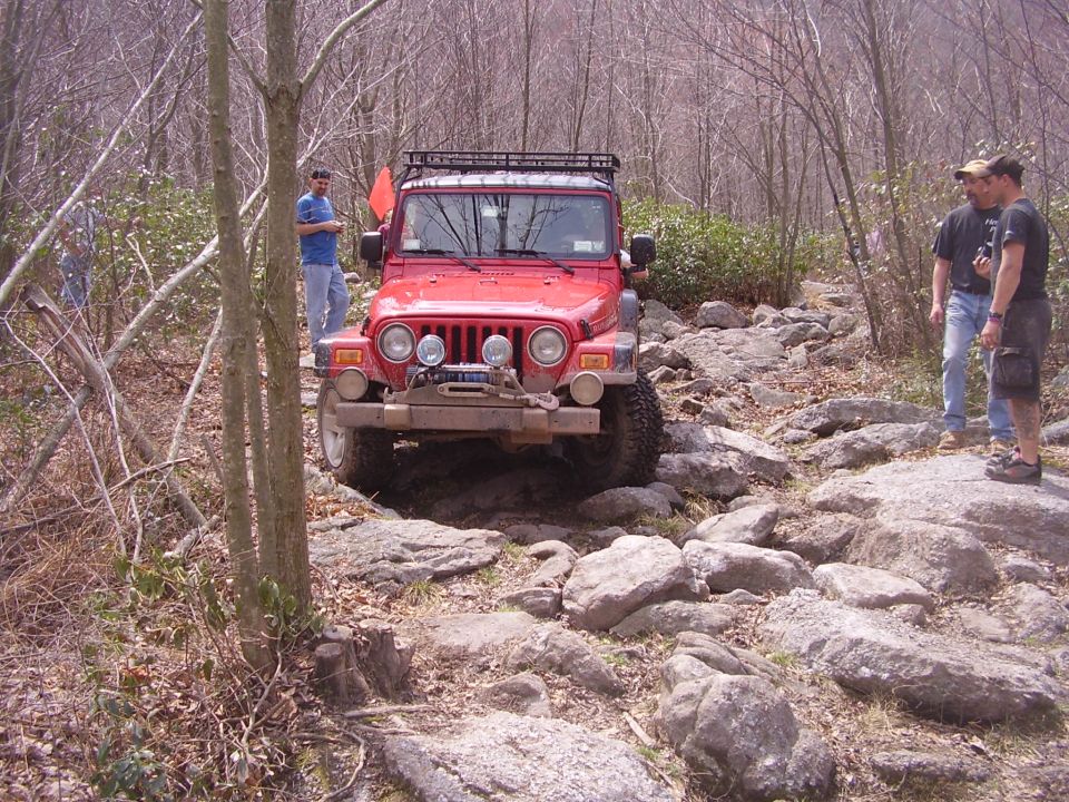 red 2003 Jeep Wrangler driving over rocks on a wooded trail