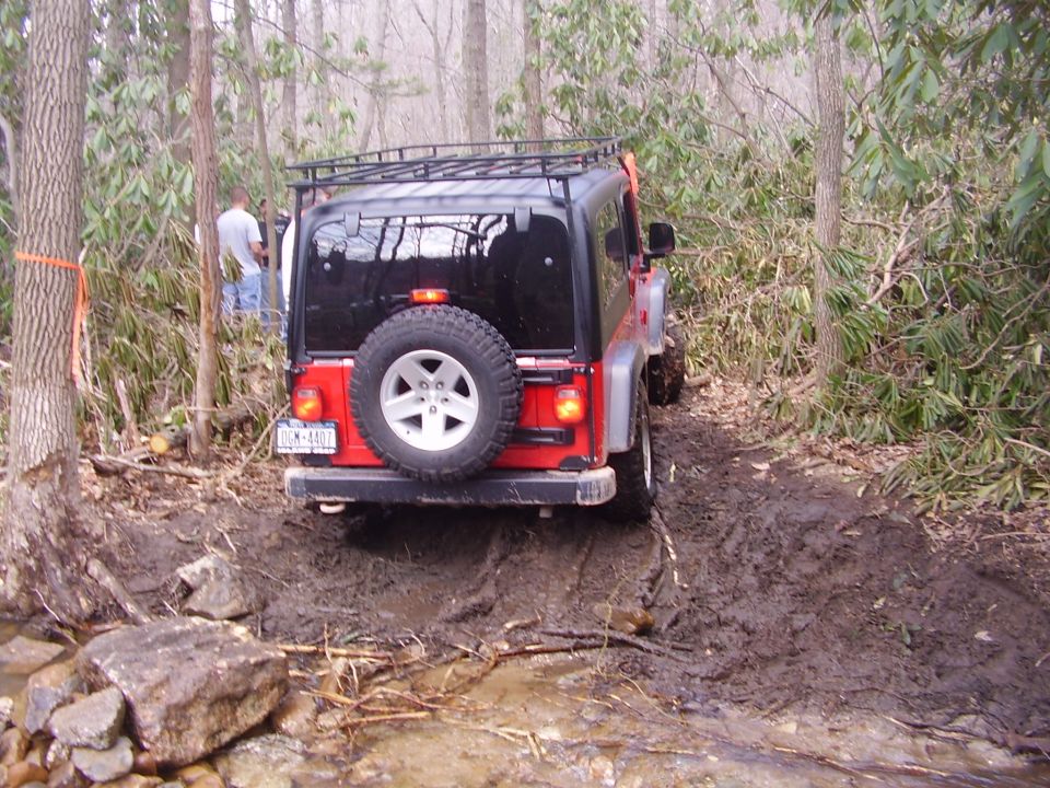 Red Jeep Wrangler with roof rack and spare tire on the back, driving on a muddy trail