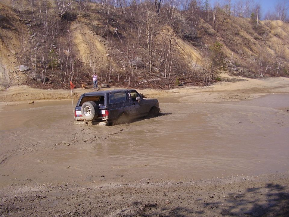 Dark SUV with spare tire on back stuck in mud pit, person standing on bank above