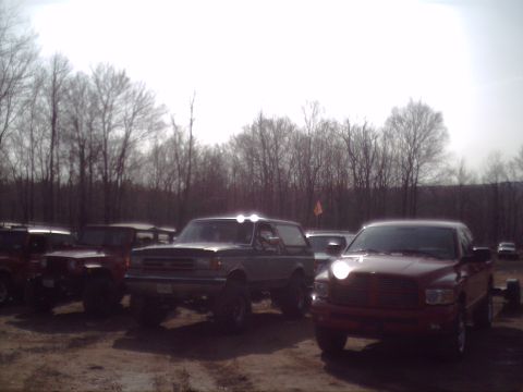 1991 Ford Bronco - Several trucks and SUVs are parked outdoors on a dirt lot with trees in the background.
