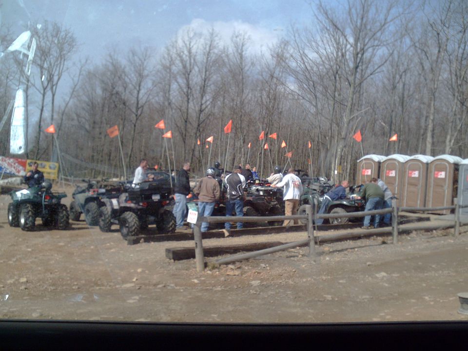 Group of people gathered around ATVs in a wooded area with portable toilets in the background