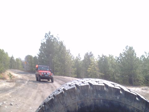 1991 Ford Bronco - A red Jeep Wrangler drives on a dirt road through a wooded area, with another vehicle's tire in the foreground.