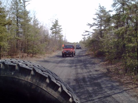1991 Ford Bronco - A convoy of Jeeps navigates a wooded dirt road, with the foreground dominated by the tire of the lead vehicle.