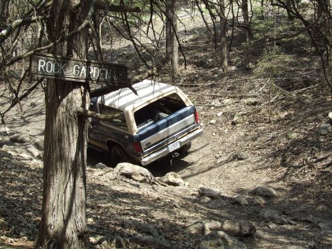1986 Ford Bronco - A Ford Bronco drives through a wooded area marked with a "Rock Garden" sign.