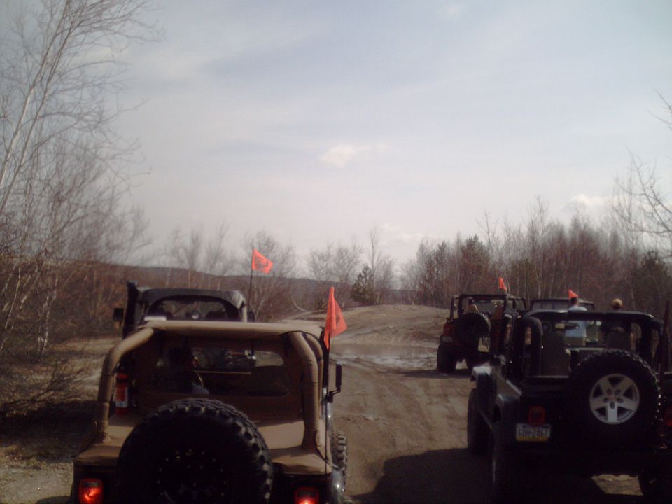 Several off-road vehicles, some with orange flags, drive on a dirt path through a wooded area