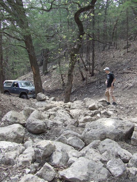 1986 Ford Bronco - A Ford Bronco drives through a rocky, wooded area, with a person observing the off-road adventure.