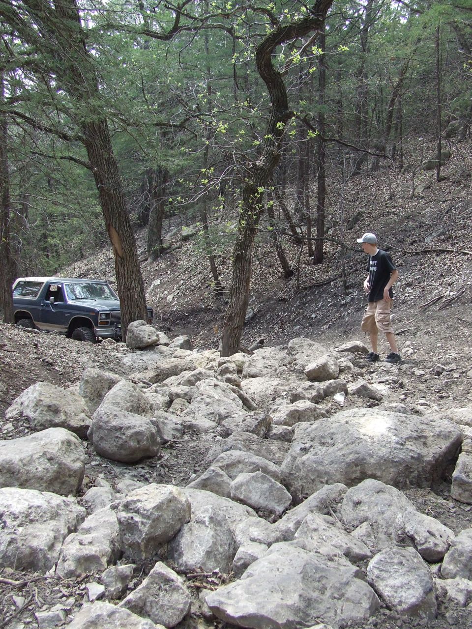 Dark blue Ford Bronco navigating a rocky, wooded trail. Person stands on the side watching