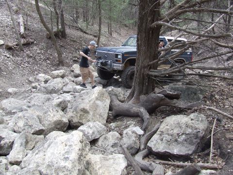 1986 Ford Bronco - A person operates a winch on a pickup truck navigating a difficult, rocky trail in a wooded area.