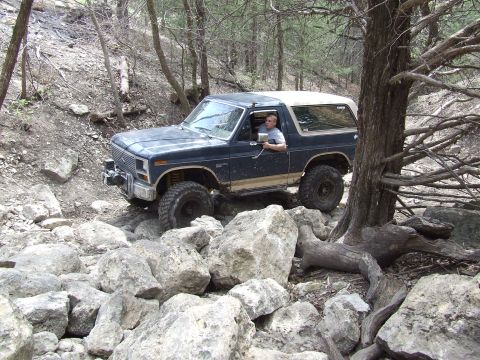 1986 Ford Bronco - A dark blue Ford Bronco with a white top is driven over a challenging rocky terrain.
