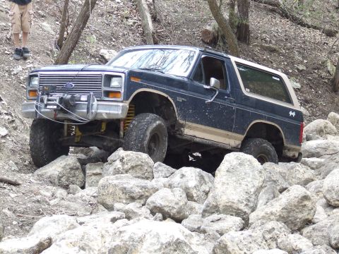 1986 Ford Bronco - This Ford Bronco is tackling a challenging off-road course with large rocks and trees.