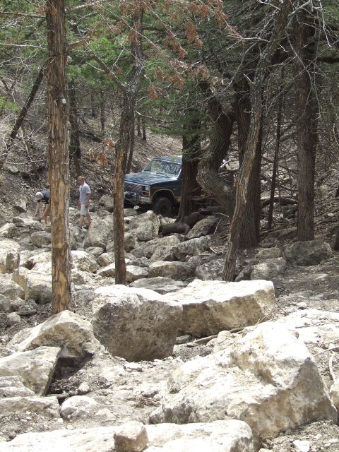 1986 Ford Bronco - A dark blue SUV drives through a challenging rocky terrain surrounded by trees.