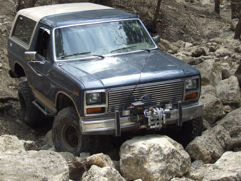 1986 Ford Bronco - This dark SUV is shown from a 3/4 front angle as it drives over rocks on a trail.