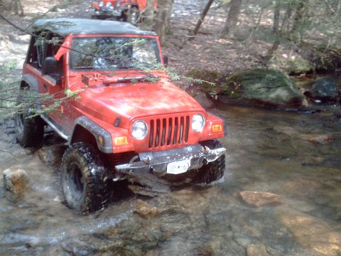 1991 Ford Bronco - Two red Jeep Wranglers navigate a shallow, rocky stream in a wooded area.