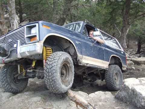 1986 Ford Bronco - A driver maneuvers a lifted Ford Bronco through a challenging, rocky outdoor terrain with trees in the background.