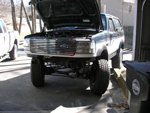 1994 Ford Bronco - The front of a lifted Ford Bronco is shown with its hood open, revealing the engine compartment and a prominent grille.