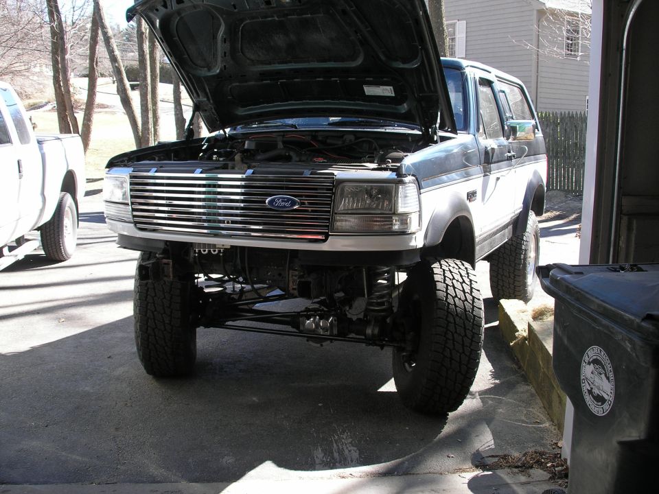 Ford Bronco, hood up, showing engine bay and front grille. Lifted suspension visible. Another white truck is parked to the left