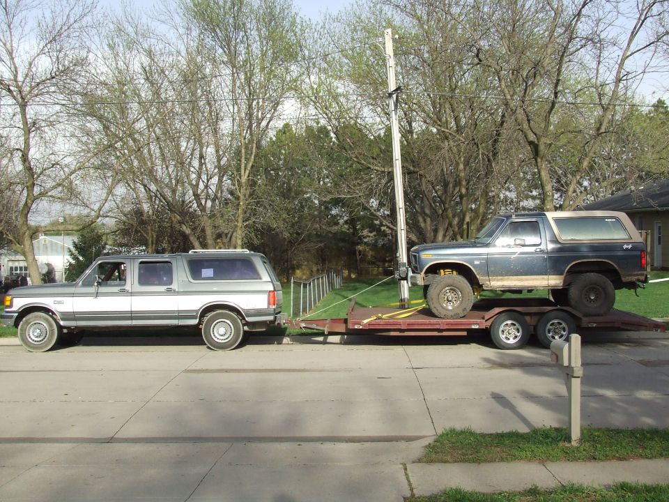 Gray two-tone Centurion crew-cab SUV towing a trailer with a lifted blue Ford Bronco on a residential street