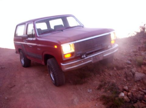 1993 Ford Bronco - A red SUV with its headlights on is parked on a dirt road with rocky terrain.
