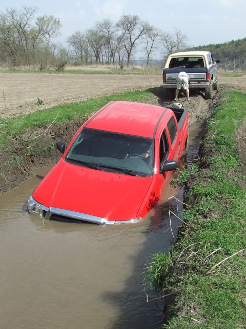 1986 Ford Bronco - Two trucks are stuck in a muddy ditch, one is a red pickup and the other is a Ford Bronco.