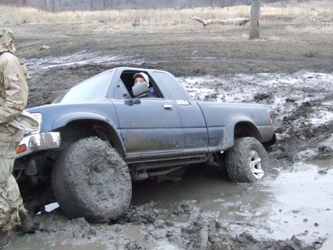 1986 Ford Bronco - A dark blue pickup truck is deeply stuck in thick mud, with a person visible in the driver's seat.