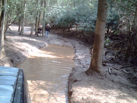 1991 Ford Bronco - A dark SUV navigates a muddy trail with water obstacles in a wooded area.