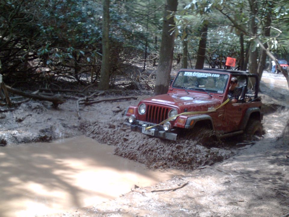 Red Jeep Wrangler stuck in a mud pit on a wooded trail. Another Jeep is visible in the background