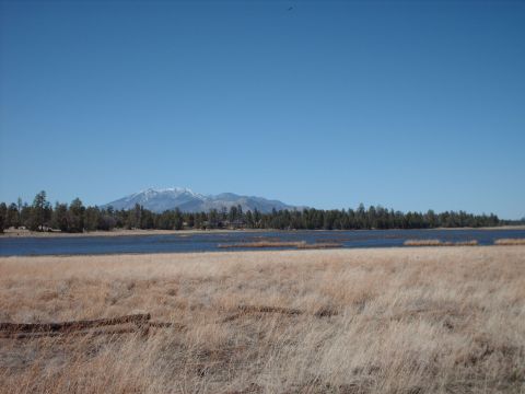 1988 Ford Bronco - A wide landscape shot shows a dry grassy field in the foreground, a calm lake, and distant snow-capped mountains under a bright blue sky.
