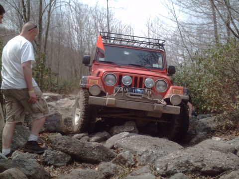 1991 Ford Bronco - A red Jeep Wrangler tackles a challenging rocky path, equipped with off-road accessories like a roof rack and winch.