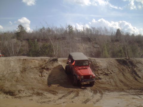 1991 Ford Bronco - Red Jeep Wrangler navigates a steep dirt incline on a sunny day with scattered clouds.