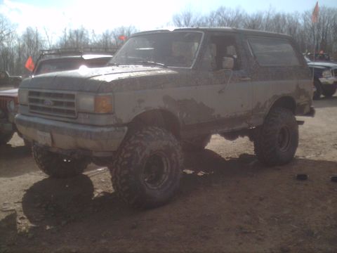 1991 Ford Bronco - A muddy Ford Bronco is parked off-road with other vehicles in the background.