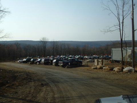 1991 Ford Bronco - A large group of SUVs and trucks are parked in a lot surrounded by trees and hills.