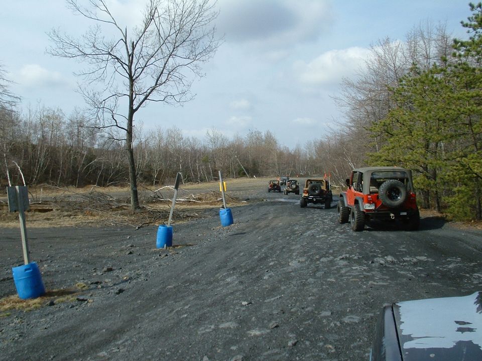 Several Jeeps driving in a line on a gravel road with trees and bare branches in the background