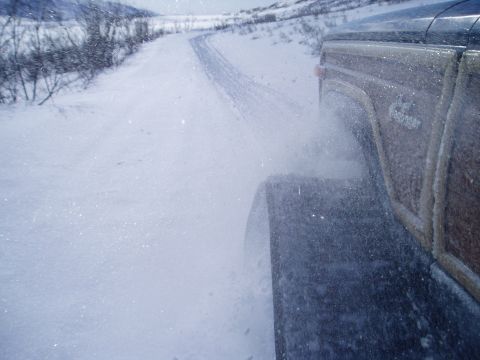 2000 Ford E350 - A wood-paneled SUV navigates a snow-covered road, with snow spraying from its tires.